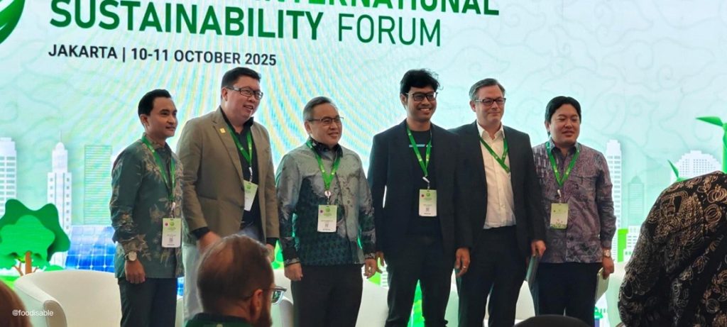 Six people wearing green lanyards stand on stage in front of a sustainability forum banner in Jakarta, posing for a photo with graphics of buildings, trees, and clouds in the background